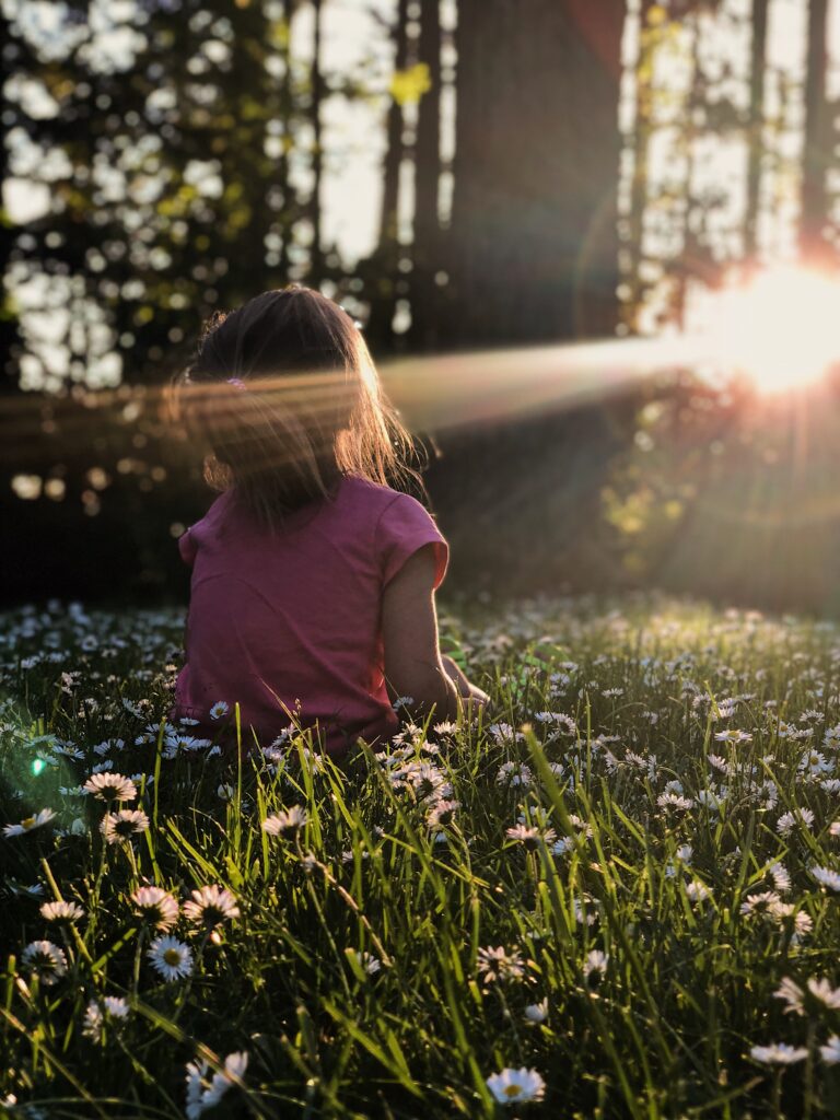 A girl sits on a woodland floor that is carpeted with daisies